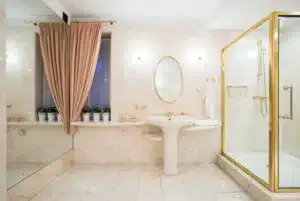 Luxurious bathroom with marble walls, gold fixtures, pedestal sink, oval mirror, glass shower, pink curtains, and potted plants on window sill.